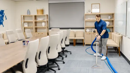 Man professionally cleaning carpet in a modern empty conference room with beige chairs and wooden shelves.