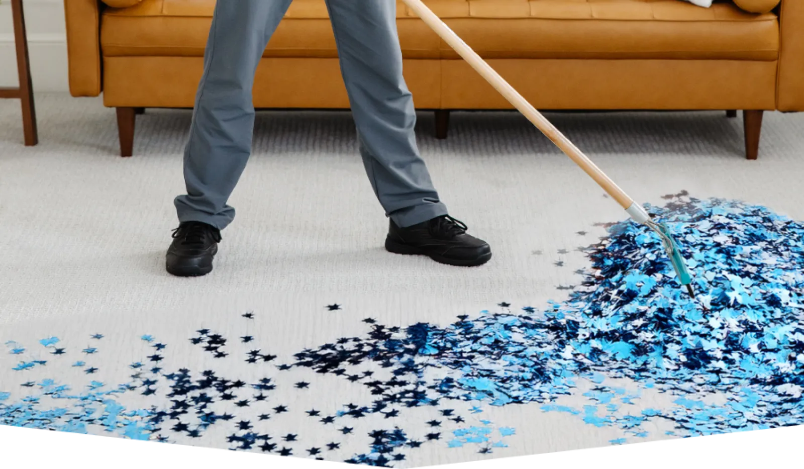 Person using a broom to sweep blue and black star-shaped confetti on a carpet near a brown sofa.